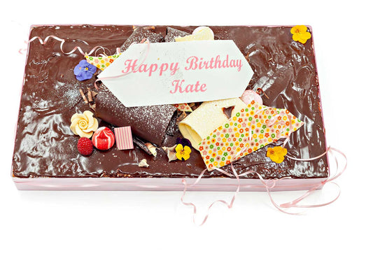 Rectangular birthday cake topped with chocolate ganache and chocolate curl decorations. It has a 'Happy Birthday Kate" inscription plaque on it, and is finished with a selection of summer flowers, summer fruit and big chocolate curls.
