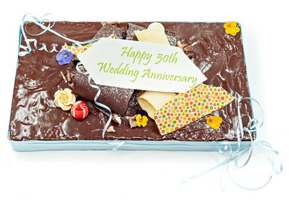 Rectangular wedding anniversary cake with a chocolate ganache on top and decorated with chocolate decorations and edible flowers.