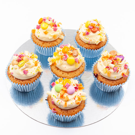 Six vanilla sponge cake cupcakes with butter cream on top, covered in a sprinking of Smarties.